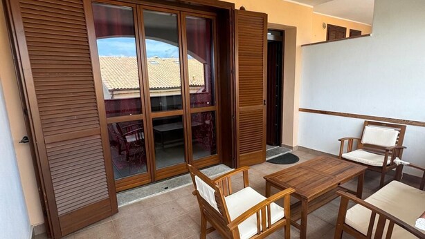 Een balkon met een houten tafel en stoelen, een schuifpui en uitzicht op een gebouw met een pannendak.