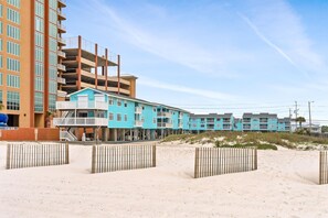 Un hotel frente a la playa con varios pisos, una playa arenosa en primer plano y un cielo despejado.