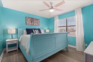 A bedroom with a bed, a nightstand, a ceiling fan, a window with blinds, and a view of the outside. - Coastal Condo Steps From the Beach (Gulf Shores)