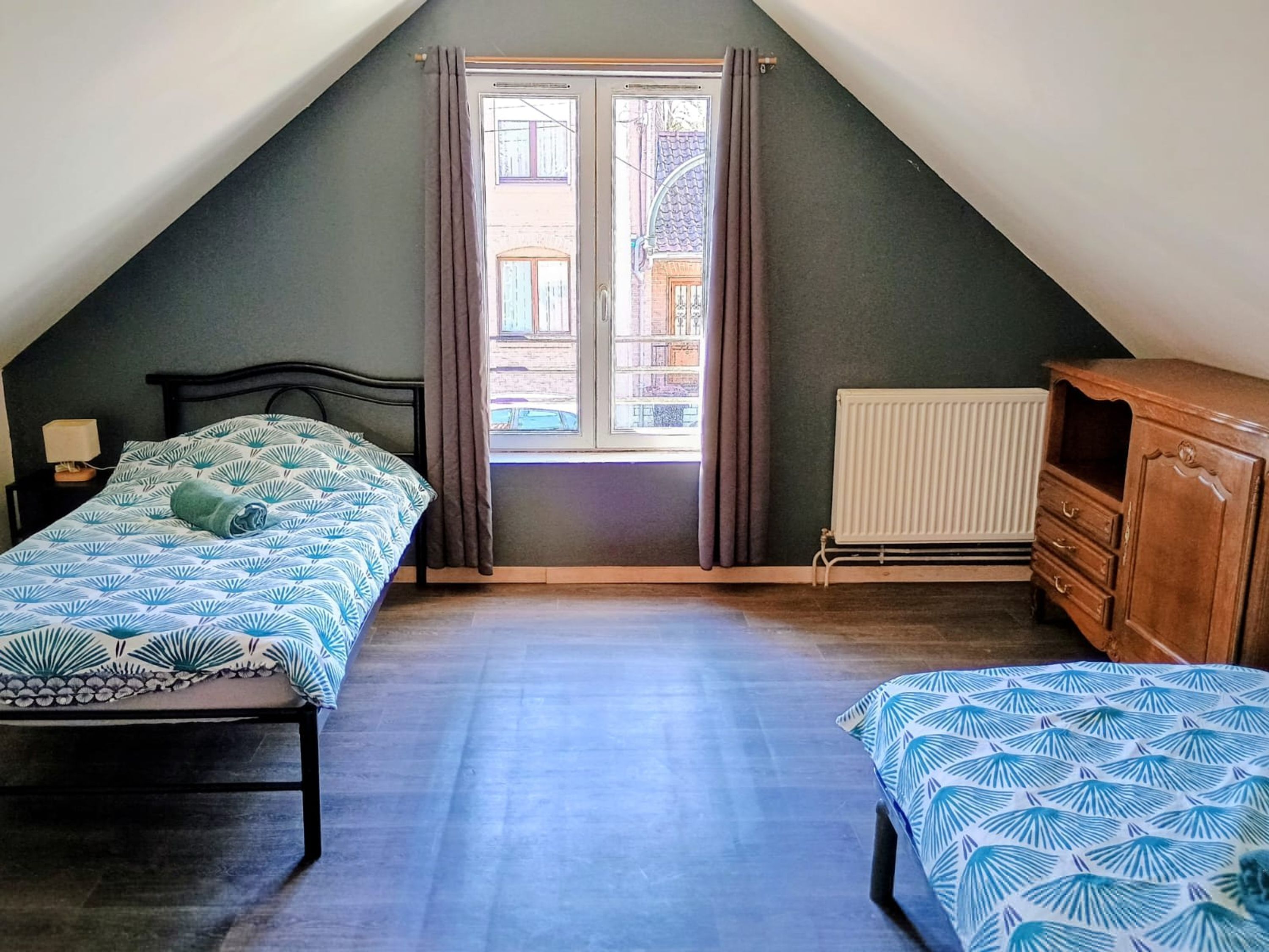 A bedroom with a sloped ceiling, a window with curtains, a radiator, a wooden dresser, and a bed with a patterned bedspread.