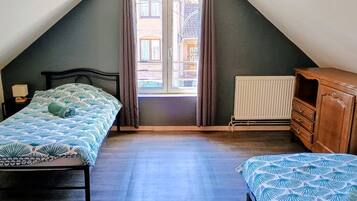 A bedroom with a sloped ceiling, a window with curtains, a radiator, a wooden dresser, and a bed with a patterned bedspread.