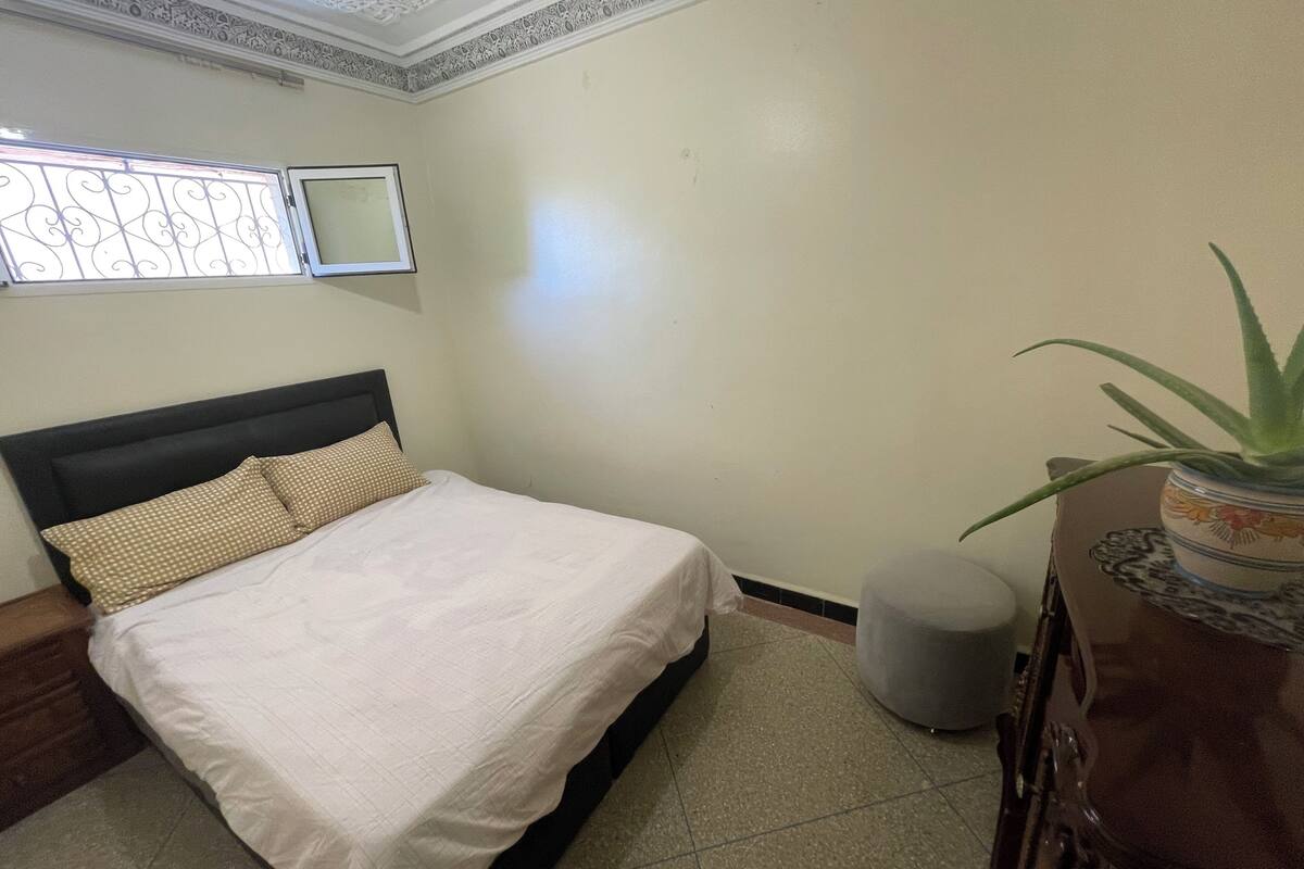 Ein Schlafzimmer mit einem Bett, einem Fenster mit Metallgitter, einer Topfpflanze und einem kleinen runden Pouf.