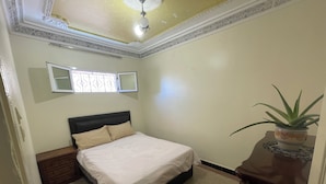 Een slaapkamer met een bed, nachtkastje, een potplant, een raam met decoratieve roede en een plafond met lamp.