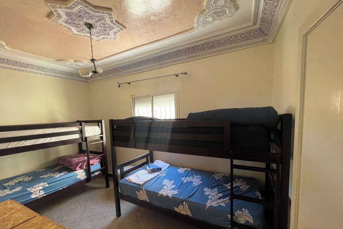 Ein Zimmer mit zwei Etagenbetten, einer Holzbank, einem Deckenventilator und einem Fenster mit Jalousien.