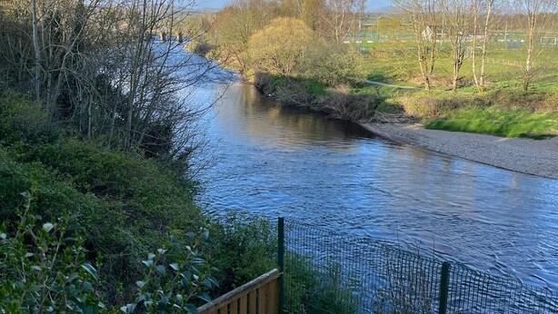 Een rivier met een houten terras en een groen gazon, omringd door bomen en een heldere lucht.