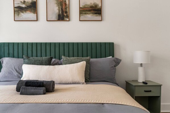 A bed with a green headboard, multiple pillows, a white throw blanket, and a bedside table with a lamp.
