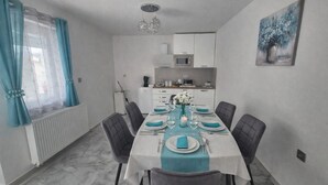 Een eethoek met een tafel voor zes personen, stoelen, een keuken op de achtergrond en een schilderij aan de muur.