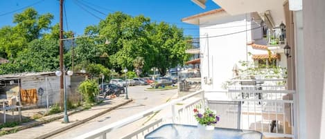 Un balcon avec une table et des chaises, donnant sur une rue bordée de voitures garées et d’immeubles.