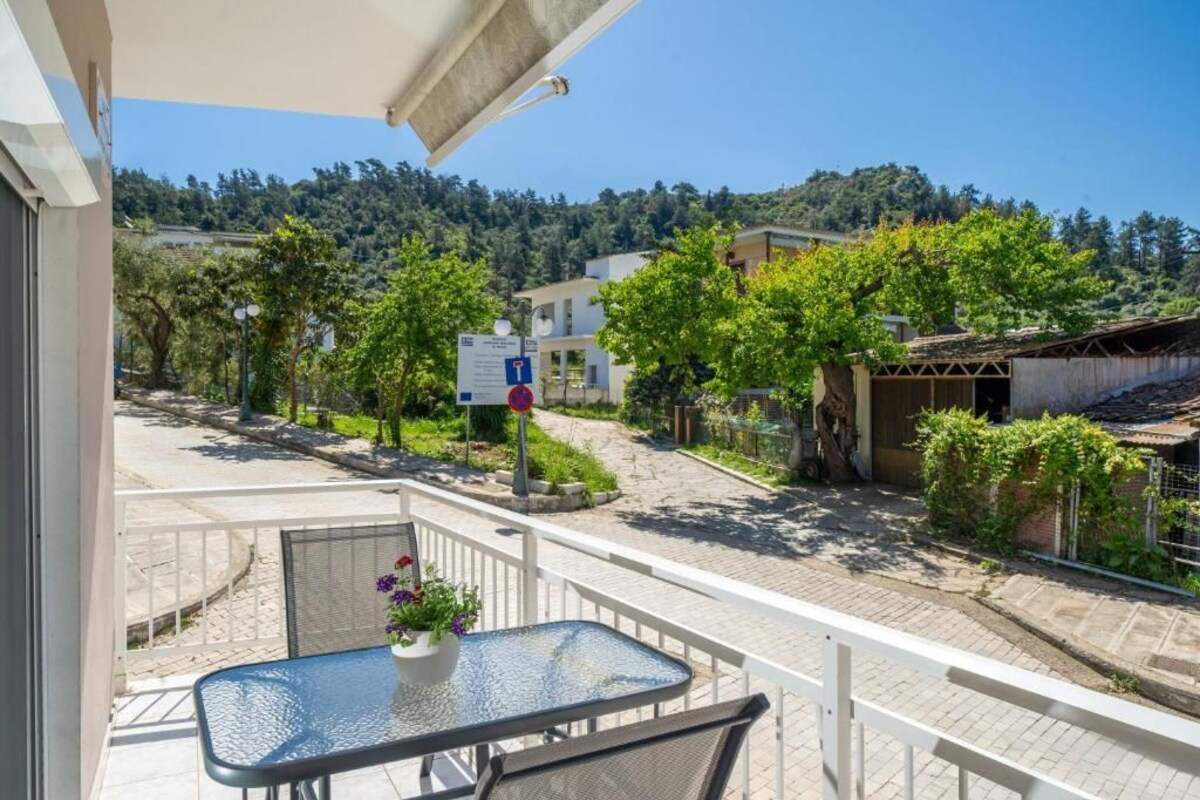 A balcony with a table and chairs, a view of a street with buildings and trees, and a clear blue sky.