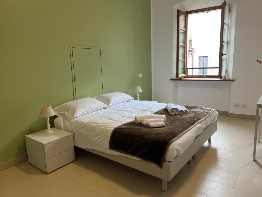 A bedroom with a bed, a nightstand, a lamp, a window, and a view of buildings outside.