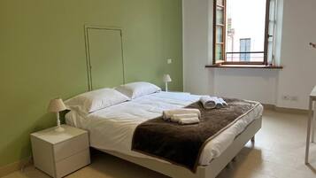 A bedroom with a bed, a nightstand, a lamp, a window, and a view of buildings outside.