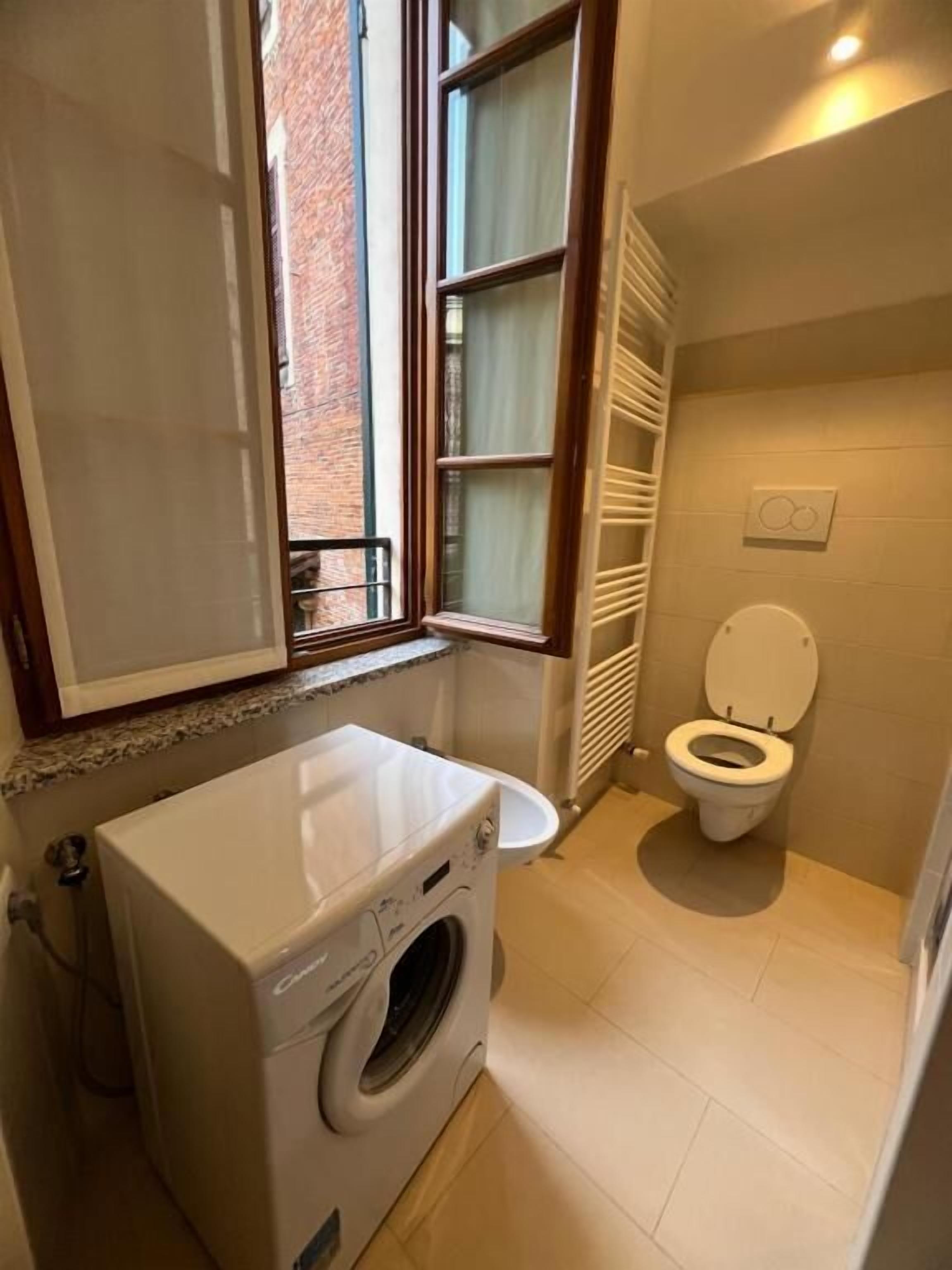 Baño pequeño con lavadora, inodoro y ventana con cortinas.