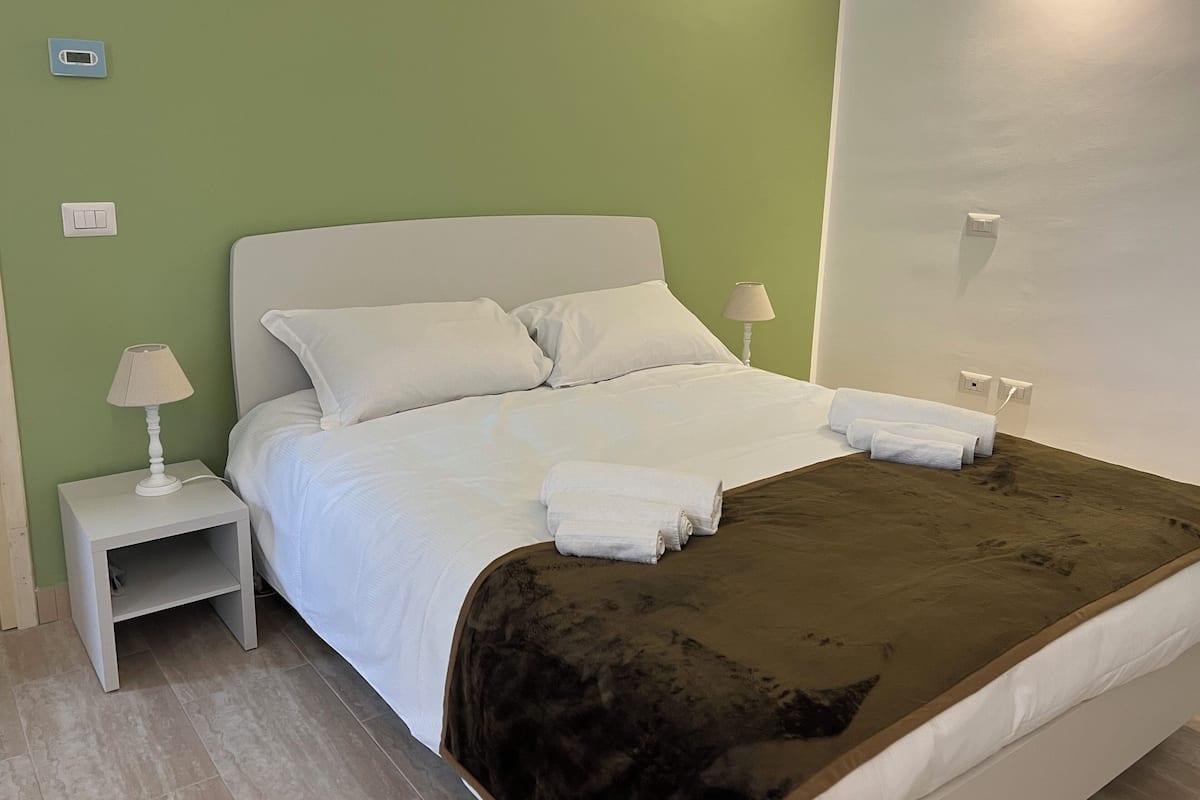 Ein ordentlich bezogenes Bett mit weißen Leinentüchern, einer braunen Tagesdecke und zwei weißen Handtüchern an der Seite. Zwei Nachttische mit Lampen und eine Steckdose an der Wand sind ebenfalls vorhanden.