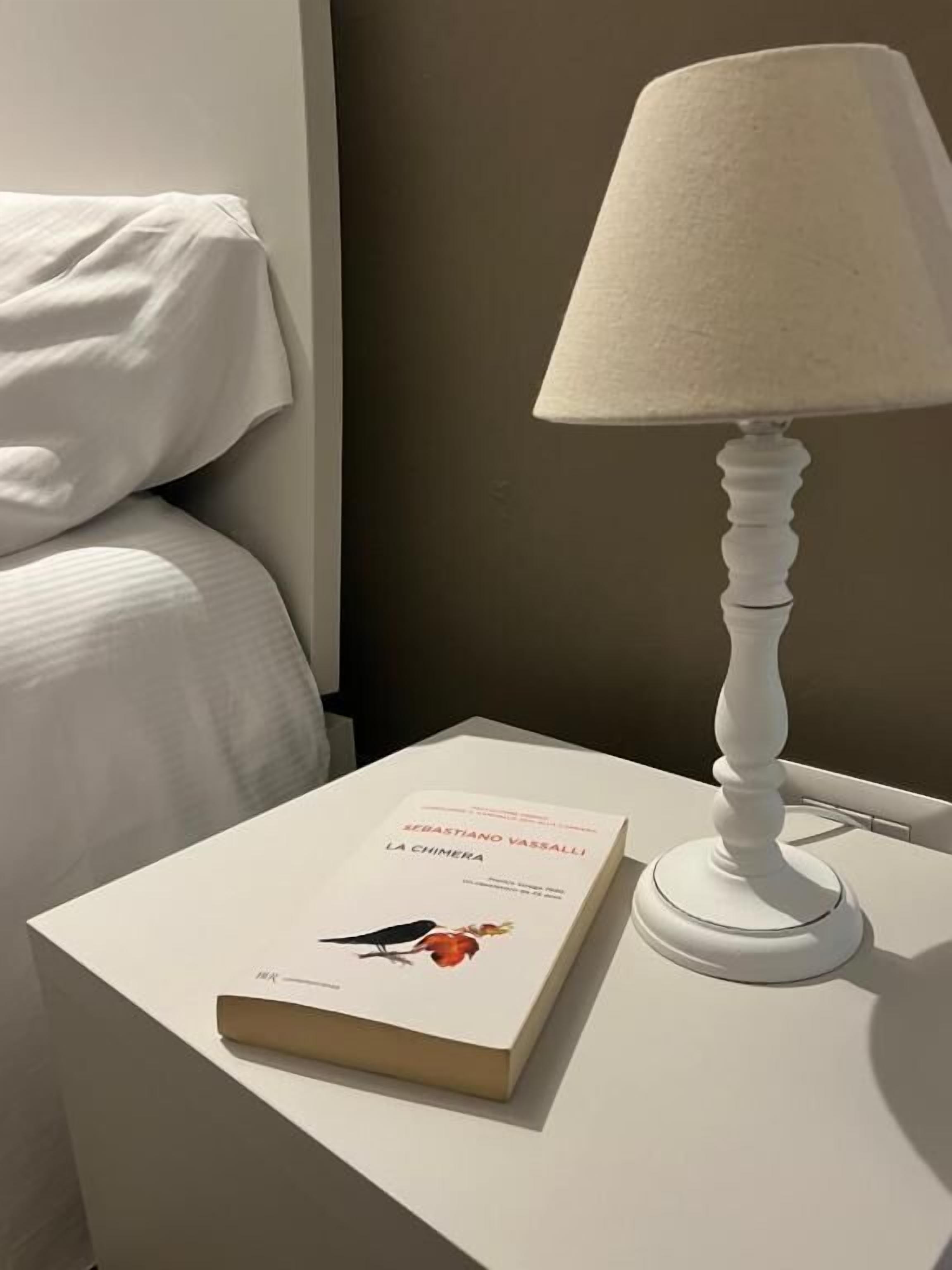Een nachtkastje met een boek getiteld 'La Chimera' van Sebastiano Vassalli, een witte lamp en een wit kussen op een bed.