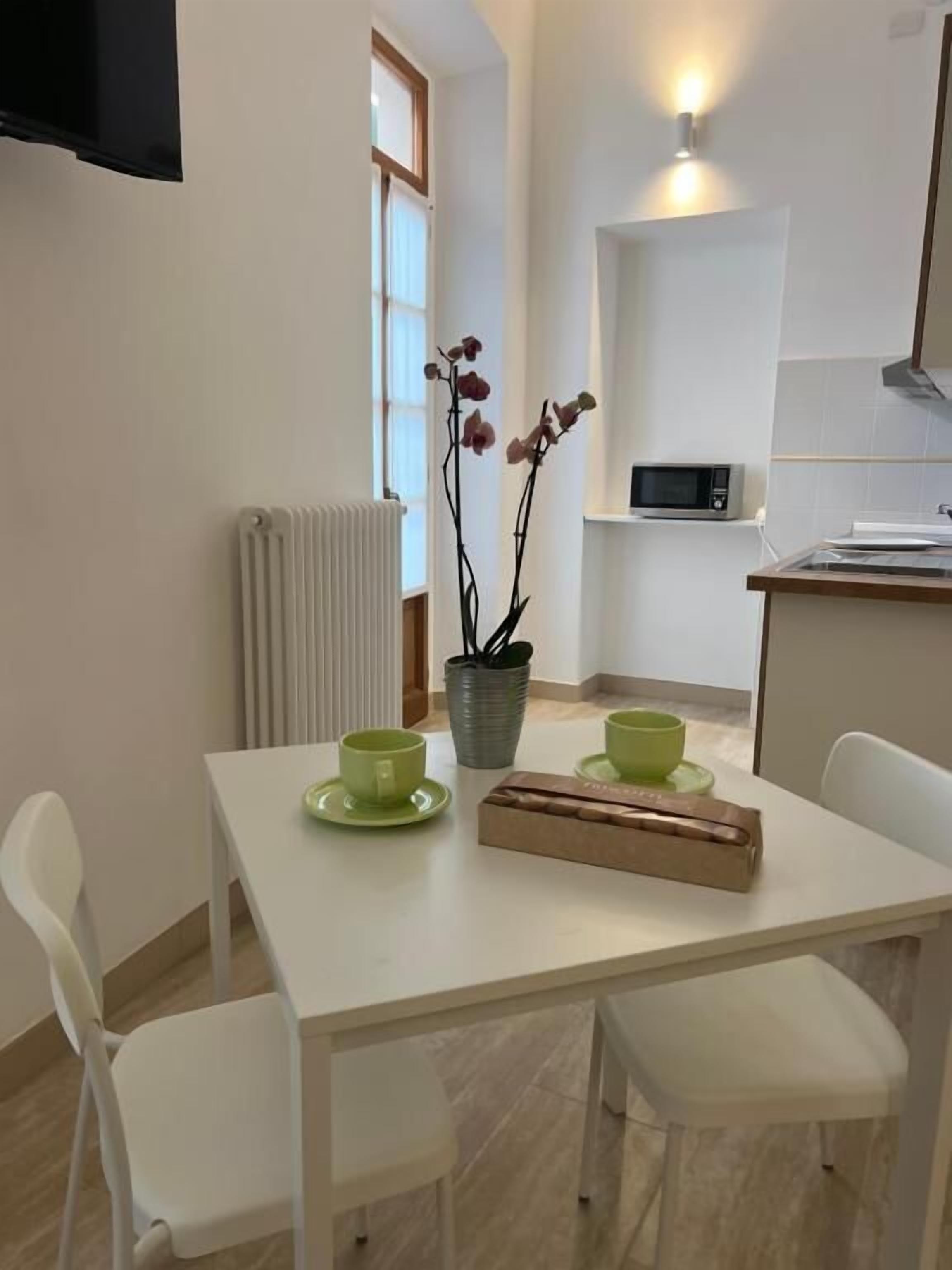 A modern kitchen with white appliances, a dining table with green cups, and a potted orchid.