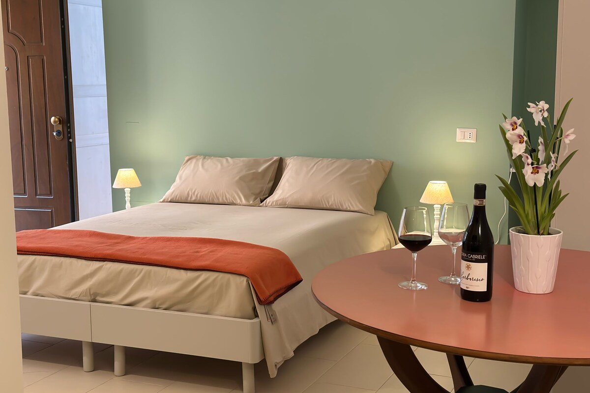 A hotel room with a bed, bedside lamps, a table with wine glasses and a bottle, and a potted orchid.