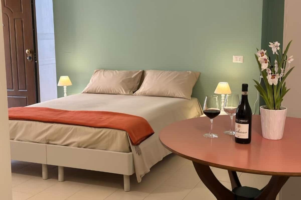 Ein Schlafzimmer mit einem Bett, Nachttischlampen, einem Tisch mit Weingläsern und einer Flasche sowie einer Topfpflanze.