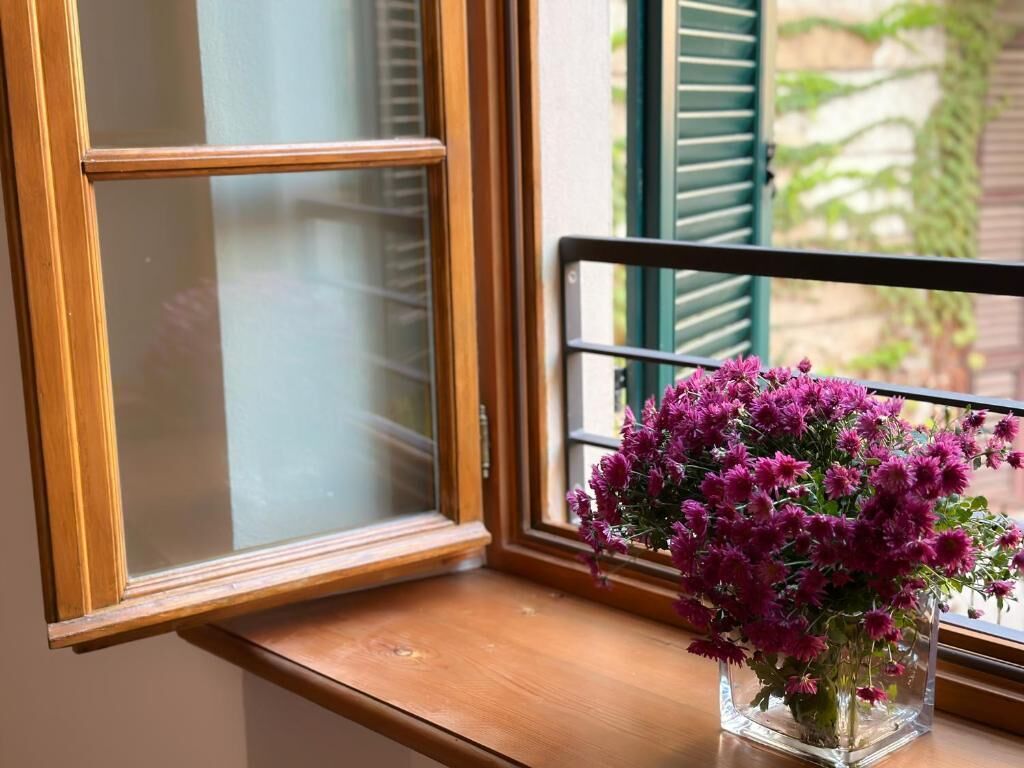 Een raam met houten frame en een heldere ruit, een houten plank eronder, en een vaas met paarse bloemen op de plank.