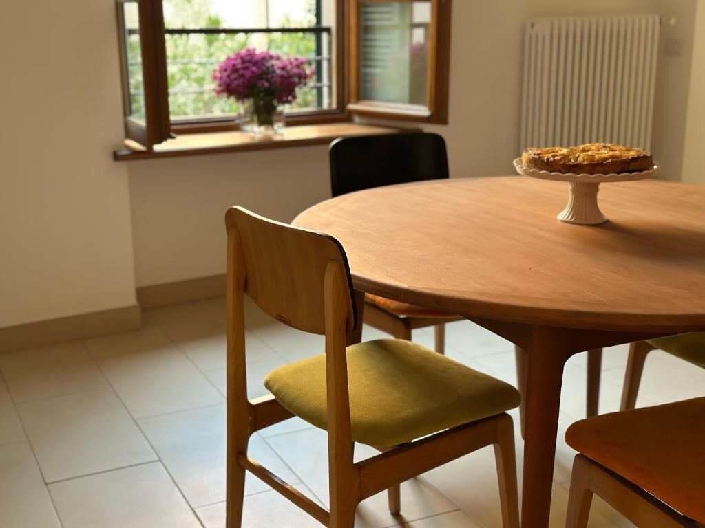 A dining table with a pie on a stand, a chair with a cushion, and a window with flowers.