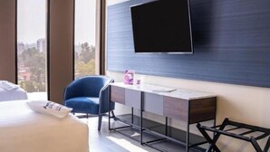 Ein modernes Hotelzimmer mit Bett, Schreibtisch, Stuhl, an der Wand montiertem Fernseher und Fenster mit Blick auf die Stadt.
