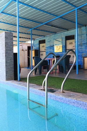 A swimming pool with metal ladders, a covered seating area with chairs, and a tiled wall with a logo. - Dahigaon Lake Resort (Karjat)