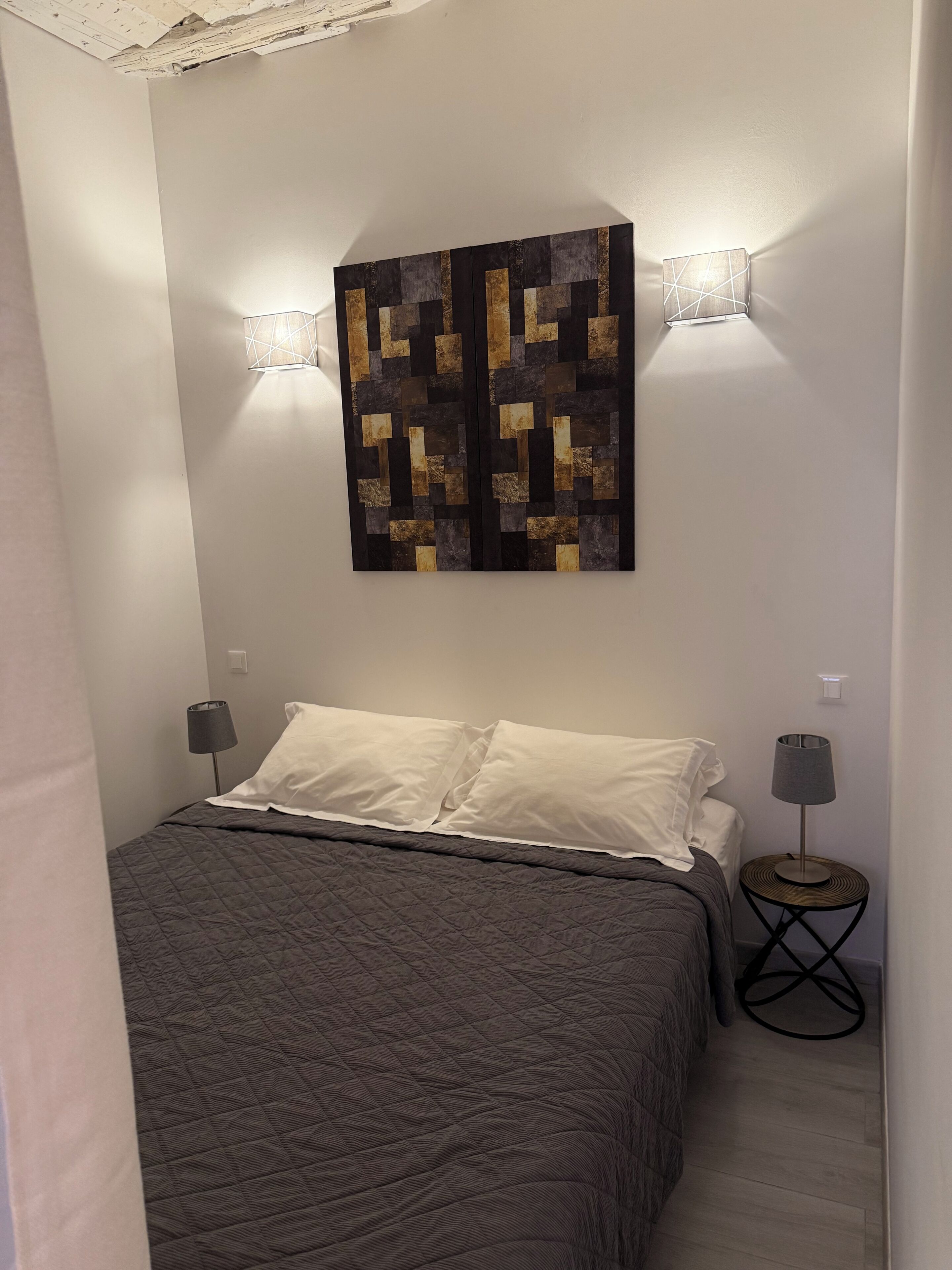 A bedroom with a bed, two bedside lamps, a wall-mounted light fixture, and a textured wall art piece.