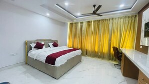 A bedroom with a bed, a desk, a chair, a ceiling fan, and a window with curtains. - sai sansthan inn (Lucknow)