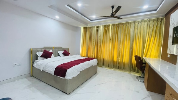A bedroom with a bed, a desk, a chair, a ceiling fan, and a window with curtains. - sai sansthan inn (Lucknow)