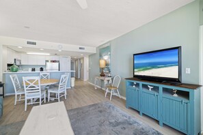 A modern kitchen with white cabinets, a dining area with a wooden table and white chairs, a television on a blue cabinet, and a view of the ocean.