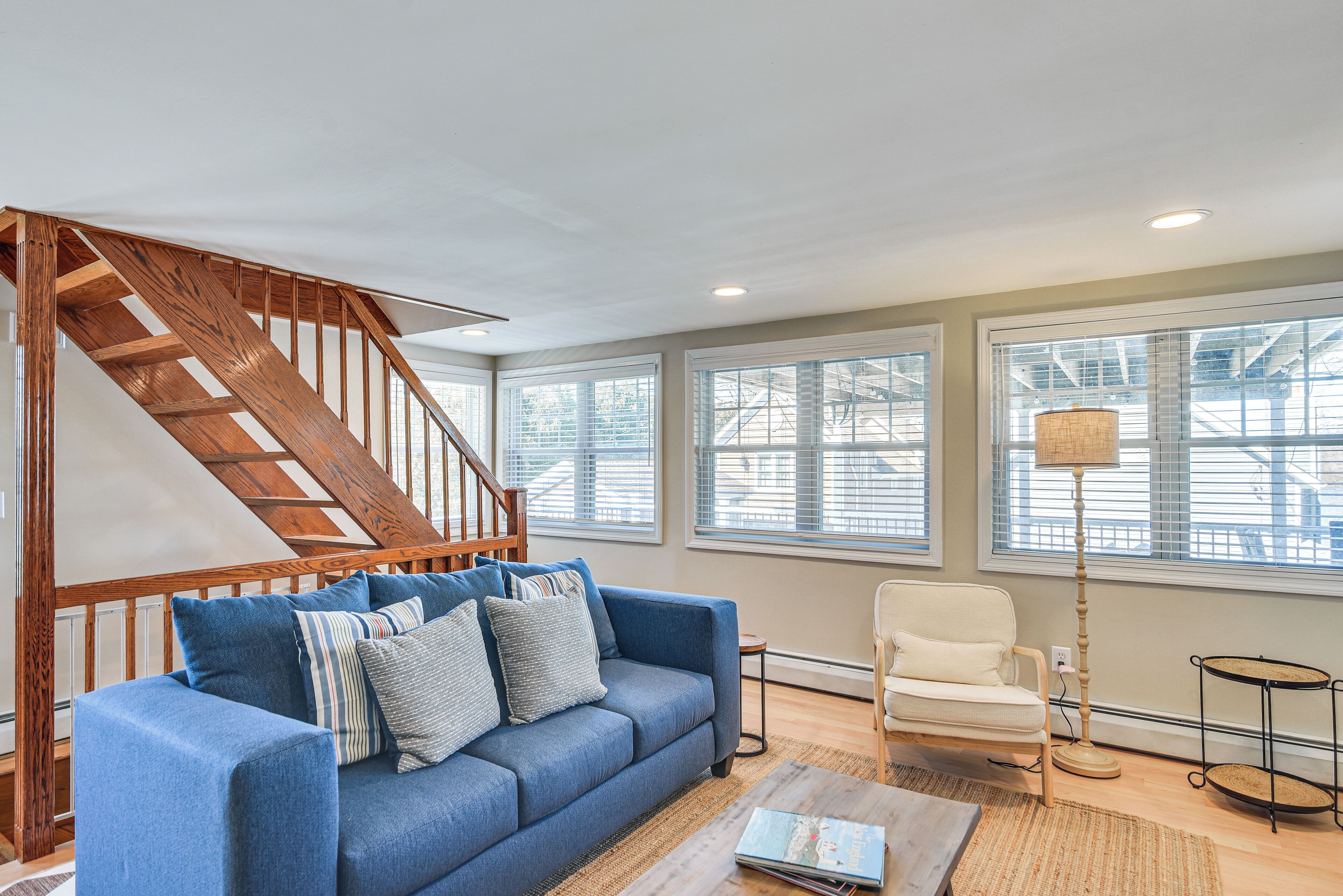 A living room with a blue sofa, a white armchair, a wooden staircase, and large windows.