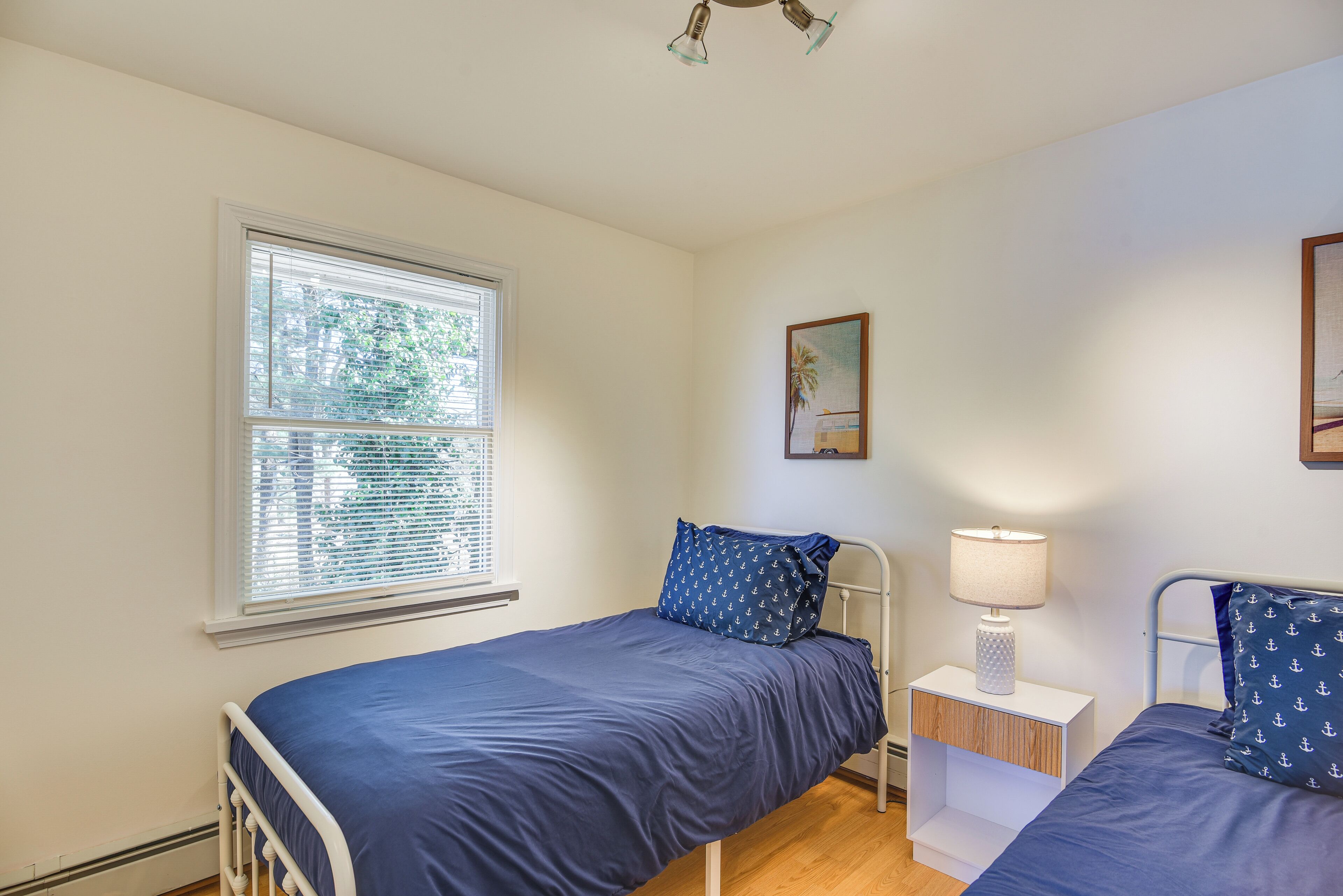 A bedroom with two beds, a window with blinds, a nightstand with a lamp, and a painting on the wall.