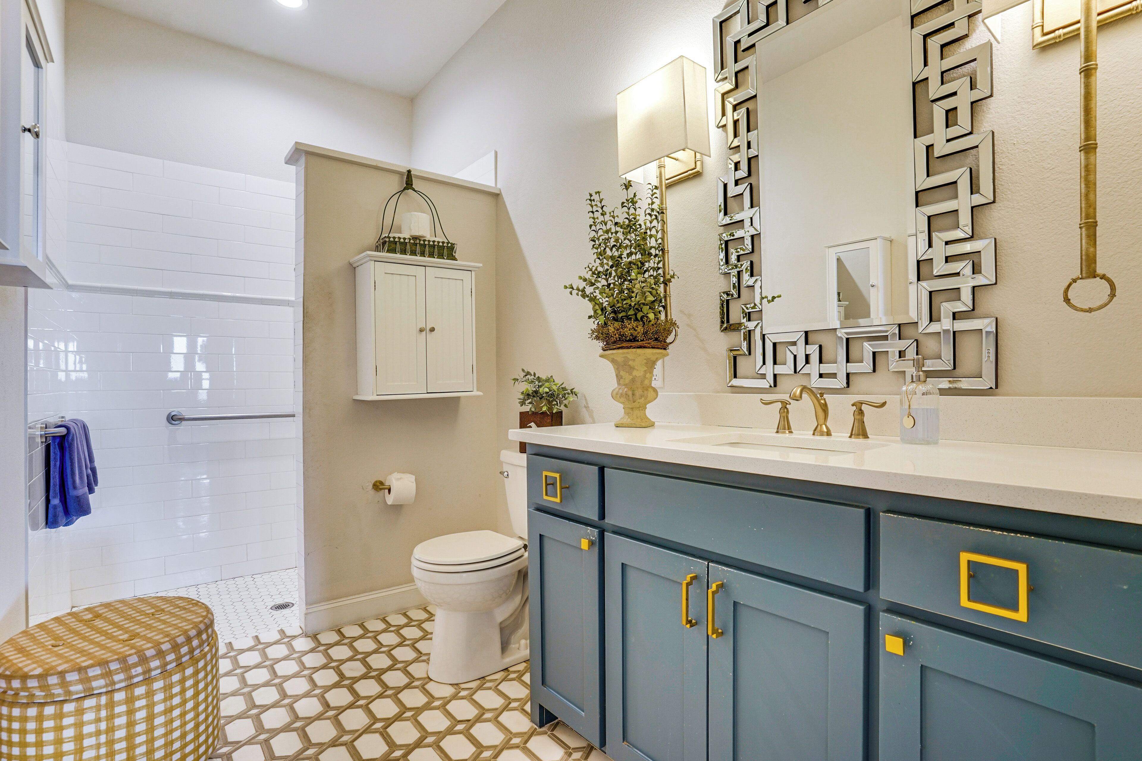 Un baño con inodoro blanco, tocador azul con pomos dorados, piso estampado y un espejo decorativo.