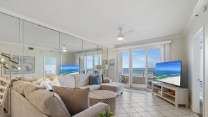 A modern living room with a large flat-screen TV, beige sofas, a round coffee table with a plant, and a view of the ocean through large windows. - The Enclave 403 by Liquid Life (Orange Beach)