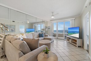 A modern living room with a large flat-screen TV, beige sofas, a round coffee table with a plant, and a view of the ocean through large windows. - The Enclave 403 by Liquid Life (Orange Beach)
