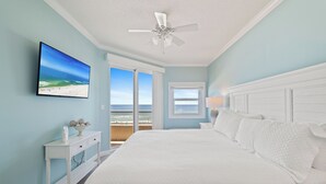 A bedroom with a bed, a ceiling fan, a TV, a nightstand, a window, and a view of the beach. - The Enclave 403 by Liquid Life (Orange Beach)