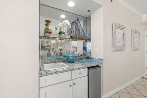 A kitchen with a large mirror, white cabinets, a stainless steel dishwasher, and a granite countertop. - The Enclave 403 by Liquid Life (Orange Beach)