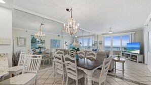 A dining area with a large table, white chairs, and a chandelier. There is a television and a balcony with a view of the ocean. - The Enclave 403 by Liquid Life (Orange Beach)