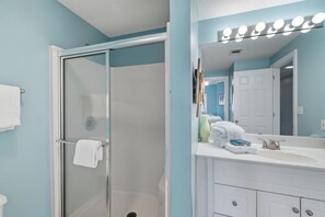 A bathroom with a glass shower door, a white sink with a mirror, and a vanity with a towel rack. - The Enclave 403 by Liquid Life (Orange Beach)