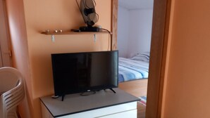 Een kamer met een televisie op een witte kast, een stoel en een bed met een gestreept laken.