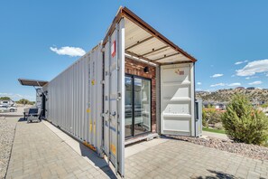 A modular building with a glass door entrance, a metal roof, and a paved area with a parked vehicle. - Mtn Views! Reimagined Container Home in Cedar City (Cedar City)