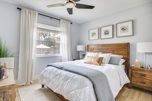A bedroom with a bed, a nightstand, a bedside table, a ceiling fan, a window with curtains, and framed pictures on the wall.