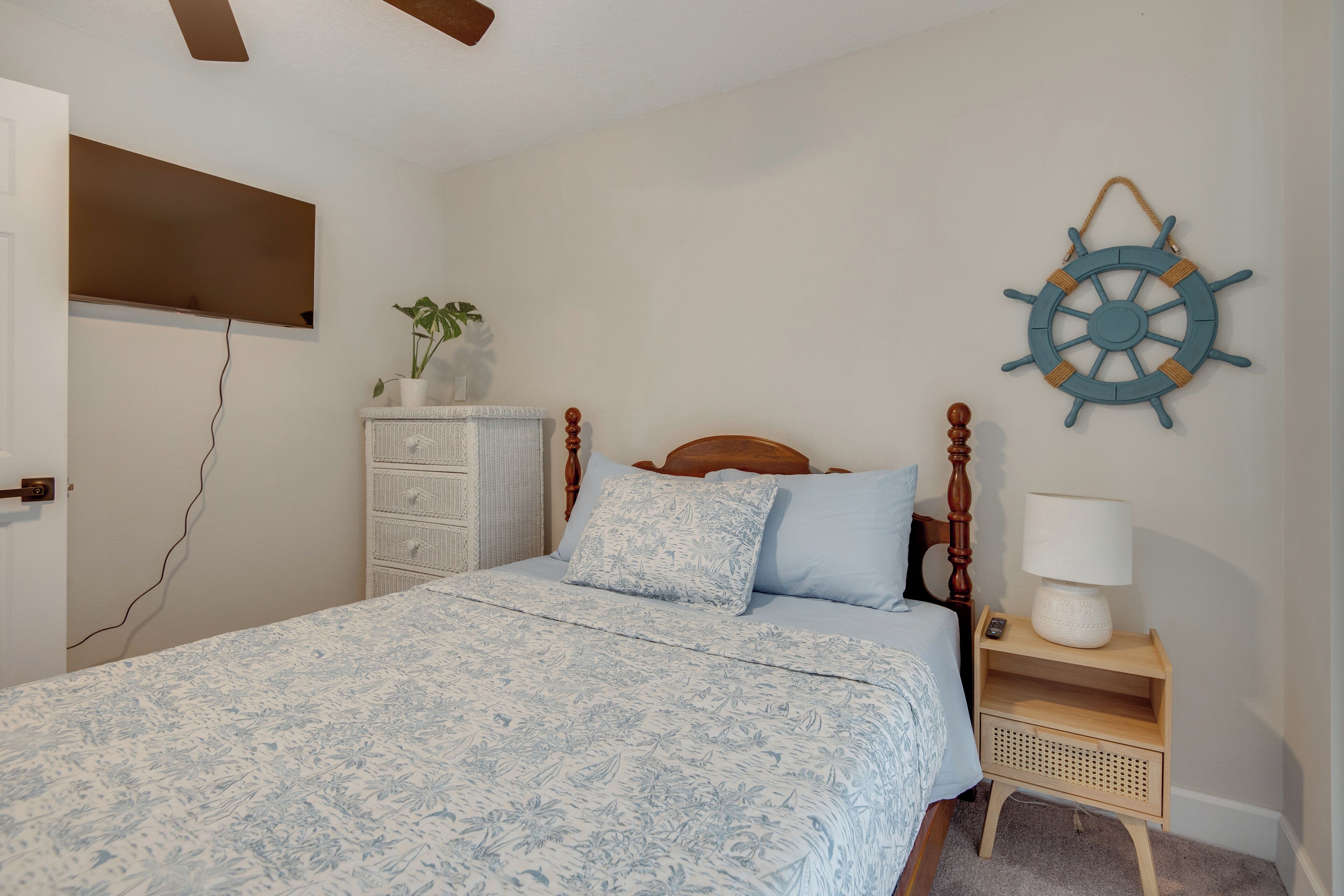 A bedroom with a bed, a nightstand, a wall-mounted ship's wheel decoration, a potted plant, and a dresser.