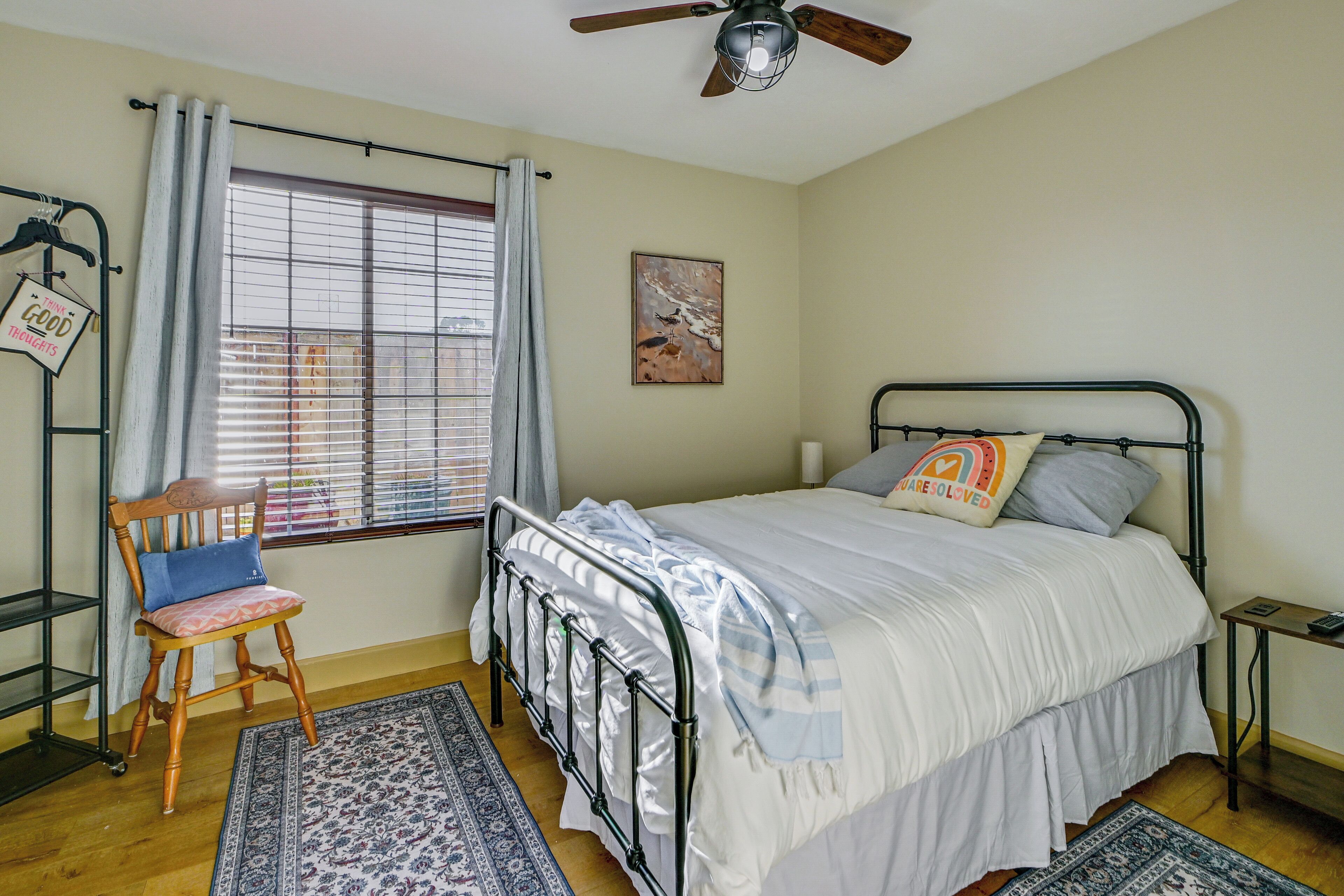 A bedroom with a bed, a chair, a window with curtains, a ceiling fan, and a wall-mounted shelf.