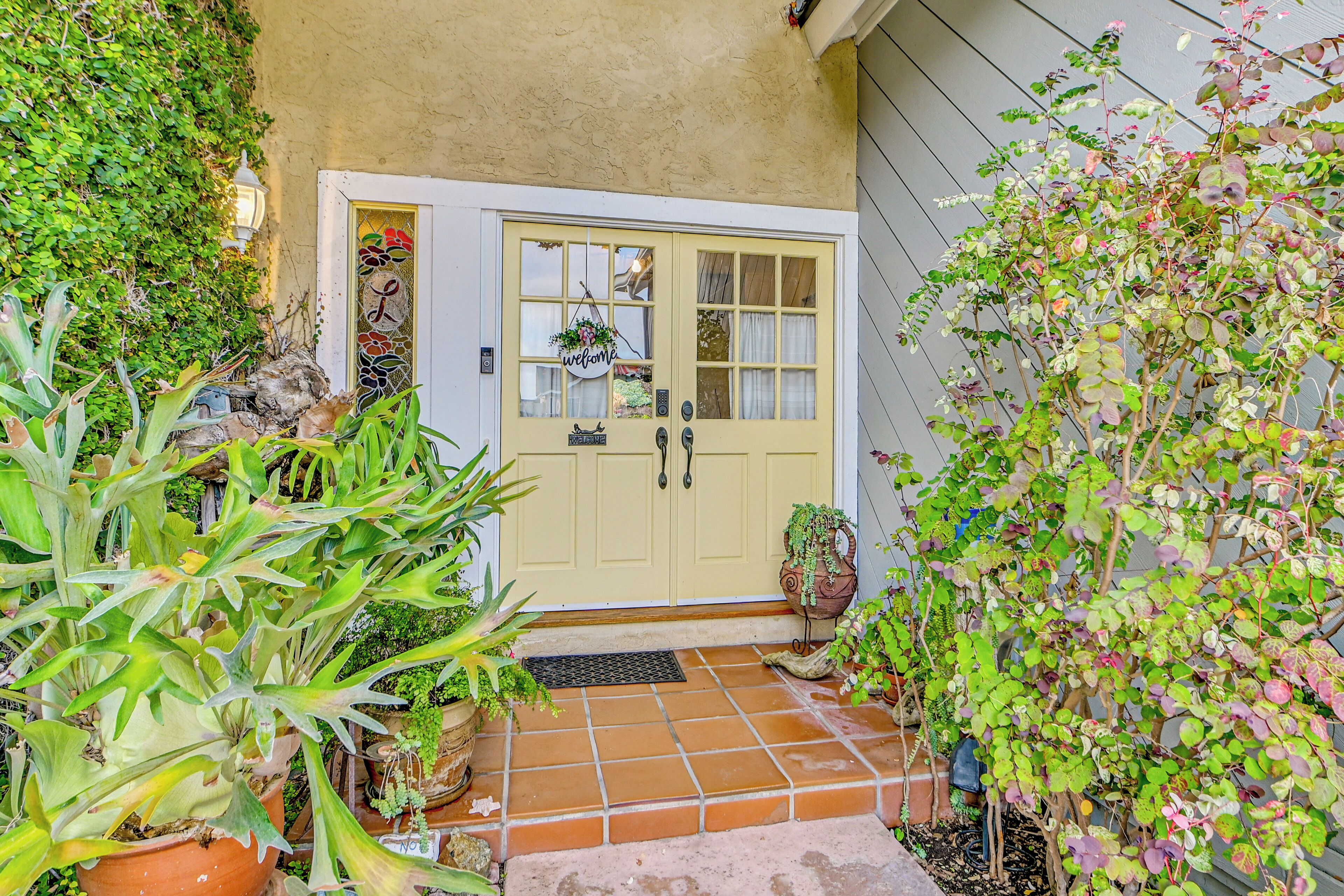 A yellow door with a 'welcome' sign, surrounded by potted plants and a tiled entrance.