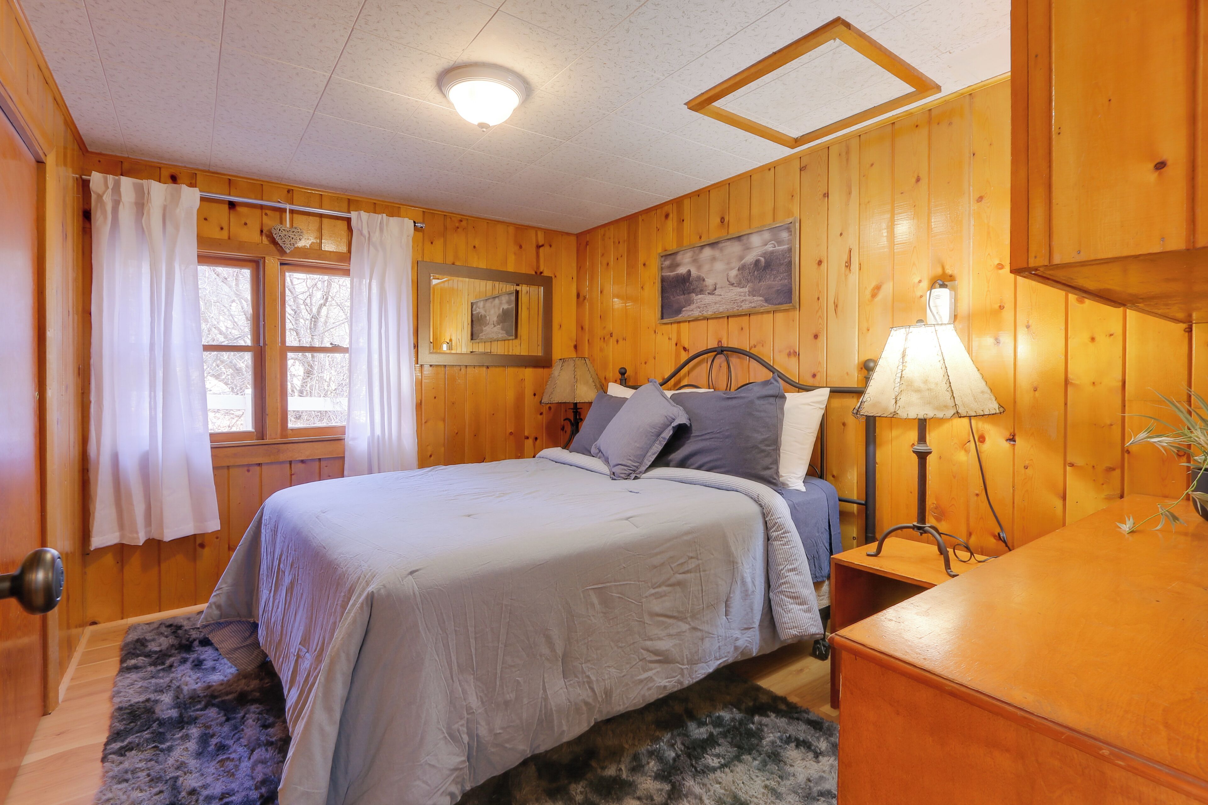 A wooden-paneled bedroom with a bed, two bedside lamps, a dresser, and a window with curtains.