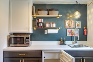 Una cocina con microondas, estantes con cestas, cafetera, tarros, un fregadero y una lámpara colgante.