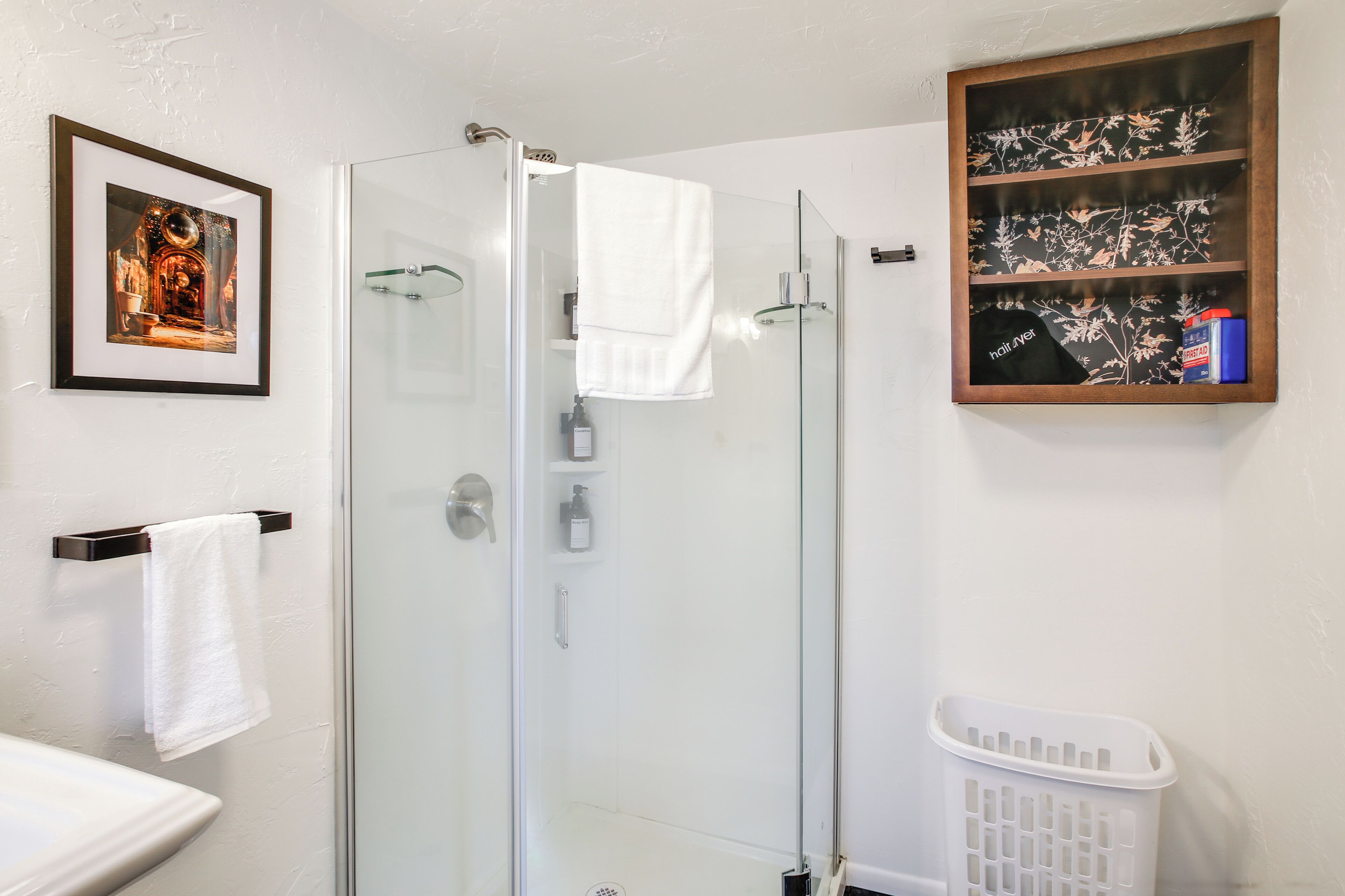 A bathroom with a glass shower enclosure, a white sink, a framed picture, a towel rack, and a laundry basket.