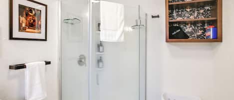 A bathroom with a glass shower enclosure, a white sink, a framed picture, a towel rack, and a laundry basket.