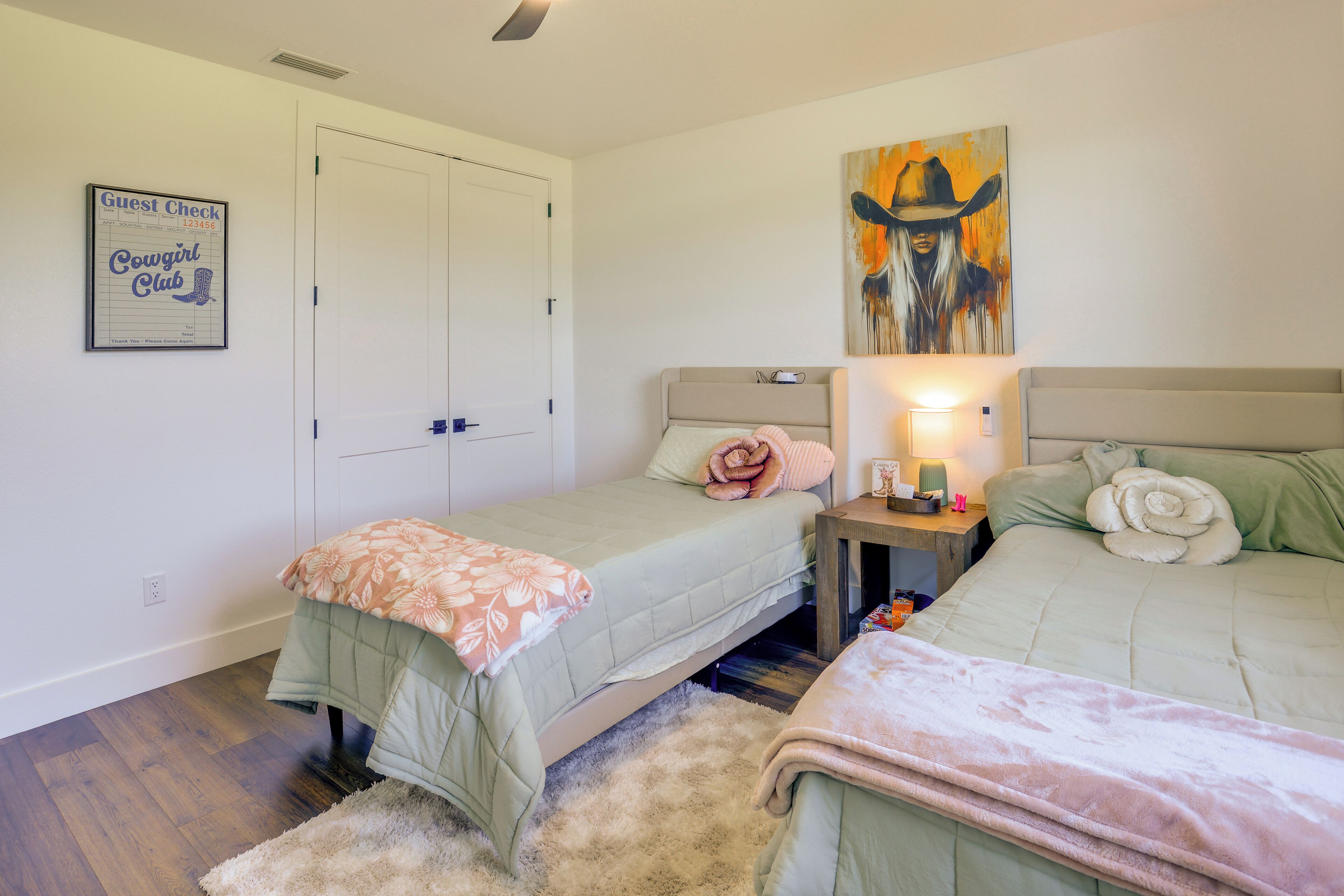 A bedroom with two beds, a nightstand with a lamp, a framed artwork, and a 'Guest Check' sign.