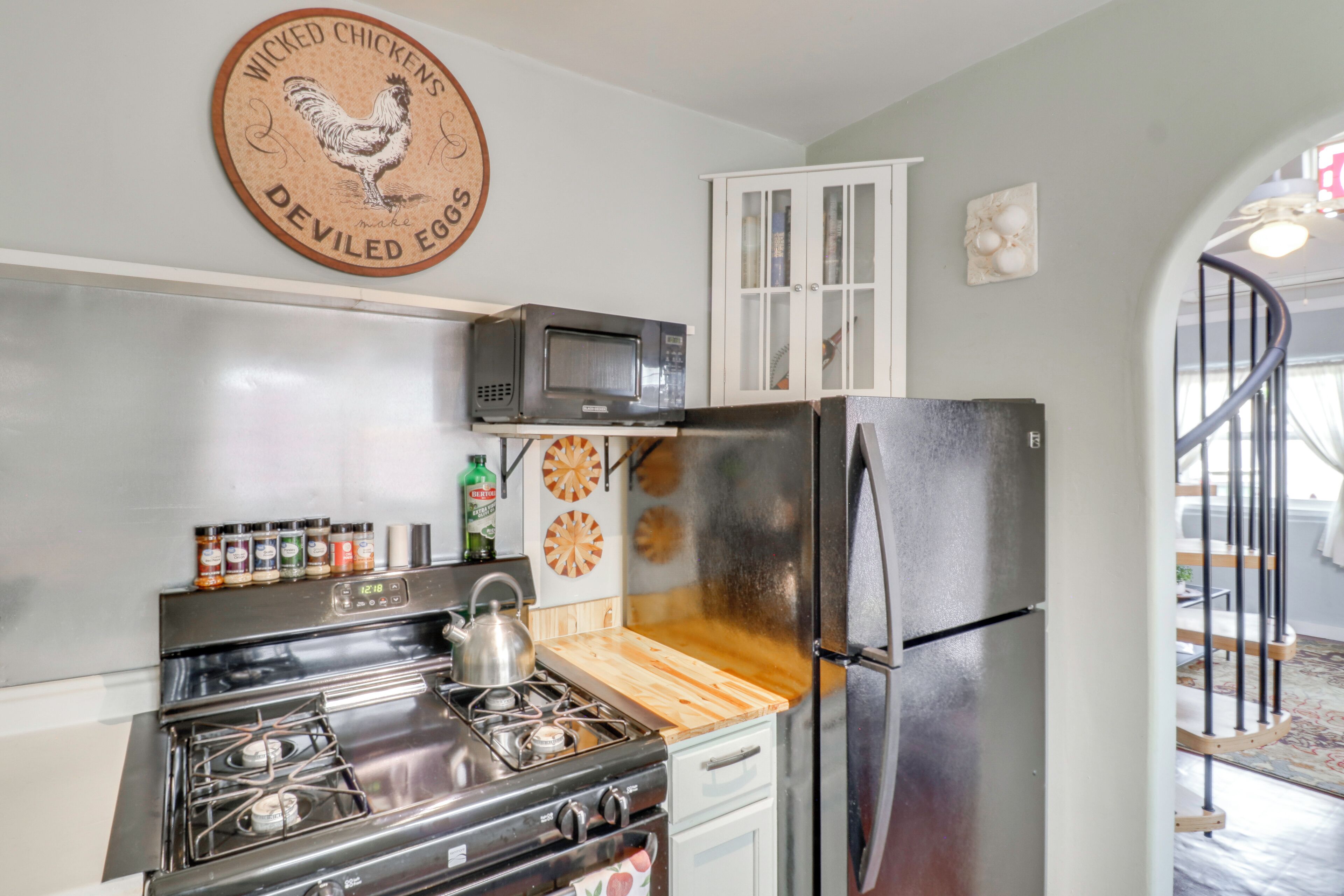Una cocina con estufa, microondas y refrigerador. Hay una tabla de cortar de madera, un armario y un reloj de pared.